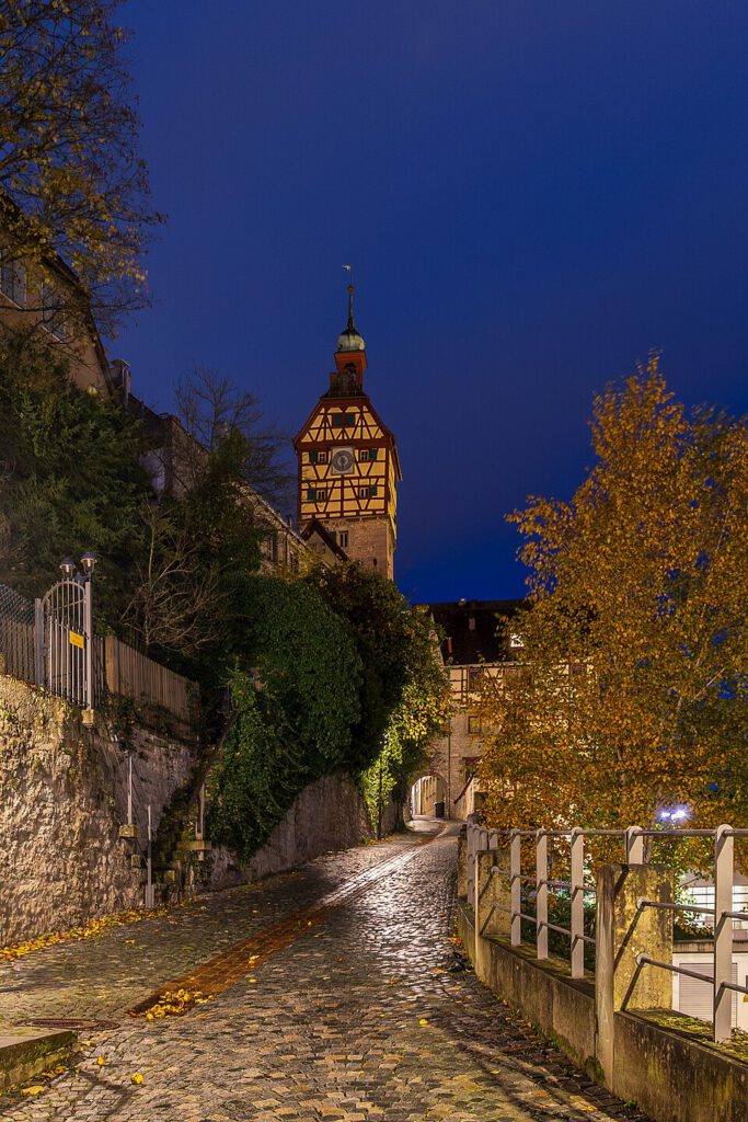 Pflasterstraße in einer historischen Stadt bei Nacht, mit einem markanten Fachwerkhaus und einem Uhrturm im Hintergrund, umgeben von Bäumen und sanften Lichtern.