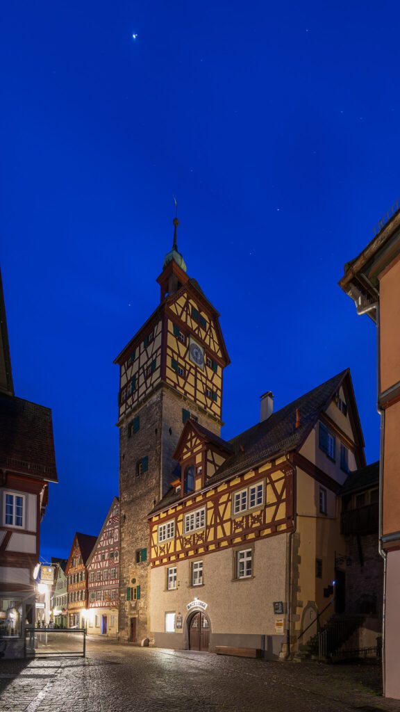 Historische Altstadt mit Fachwerkhäusern und dem markanten Uhrturm bei Nacht, unter einem klaren blauen Himmel.