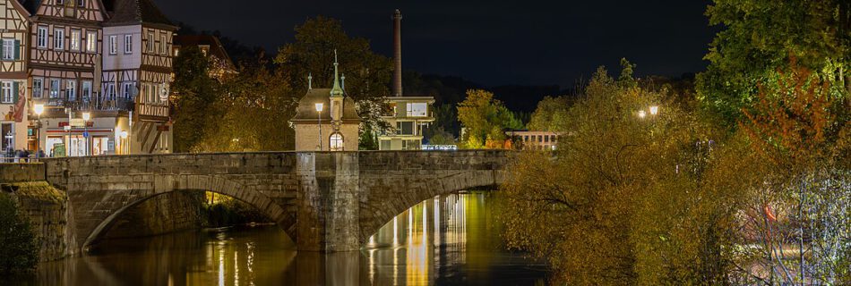 Nachtansicht der Steinbrücke in einer malerischen Stadt, umgeben von historischen Fachwerkhäusern und beleuchteten Bäumen am Ufer. Die ruhige Wasseroberfläche reflektiert die Lichter der Umgebung.