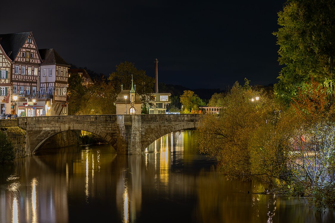 Nachtansicht der Steinbrücke in einer malerischen Stadt, umgeben von historischen Fachwerkhäusern und beleuchteten Bäumen am Ufer. Die ruhige Wasseroberfläche reflektiert die Lichter der Umgebung.
