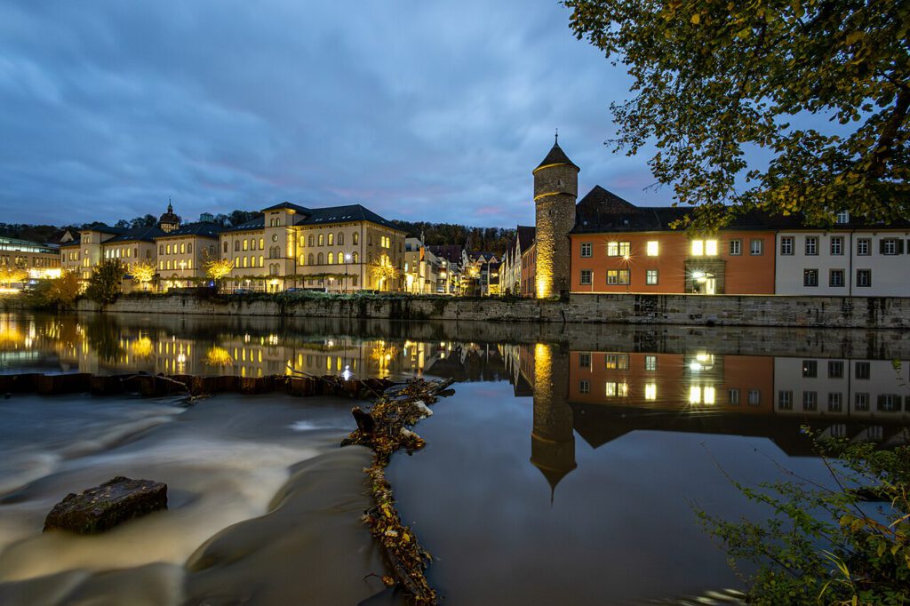 Stadtansicht bei Nacht mit beleuchteten Gebäuden entlang eines ruhigen Flusses, reflektiert im Wasser, umgeben von Bäumen und einer historischen Turmstruktur.