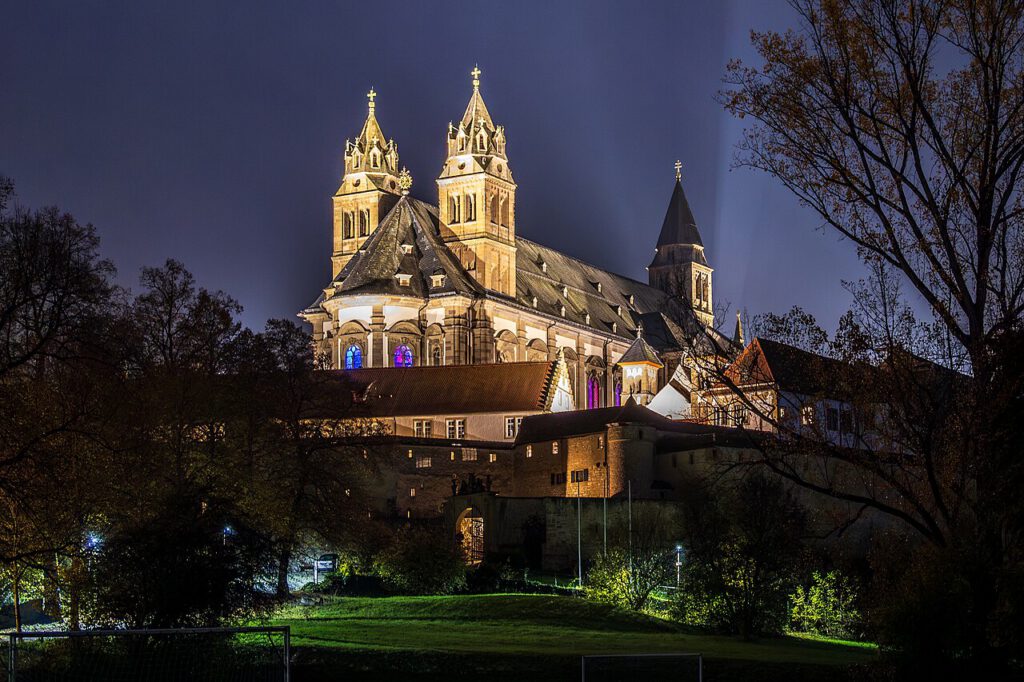 Die beleuchtete Stiftskirche mit ihren markanten Türmen und bunten Fenstern, umgeben von Bäumen und einem Park, zeigt eine beeindruckende Architektur bei Nacht.