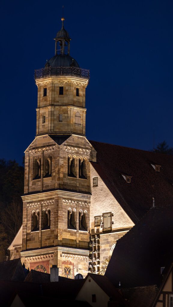 Historische Kirche mit beleuchtetem Turm bei Nacht, zeigt gotische Architektur und detaillierte Steinmetzarbeiten. Die Uhr und das Wappen sind deutlich sichtbar.