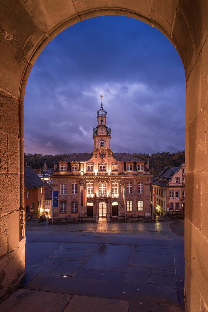 Blick durch einen mittelalterlichen Torbogen auf das beleuchtete Rathaus in einer malerischen Stadt bei Dämmerung, umgeben von historischen Gebäuden und sanften Hügeln.