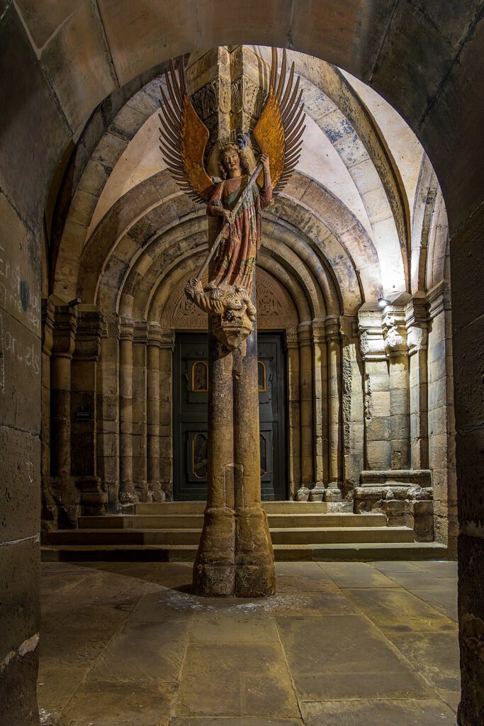 Detailreiche Darstellung eines Engelstandbildes in einem historischen Kirchenraum mit gewölbten Decken und Säulen. Der Engel mit Flügeln hält ein Schwert und steht auf einem Sockel, während im Hintergrund eine kunstvoll gestaltete Eingangstür sichtbar ist. Die Beleuchtung betont die architektonischen Elemente und die Texturen der Wände.