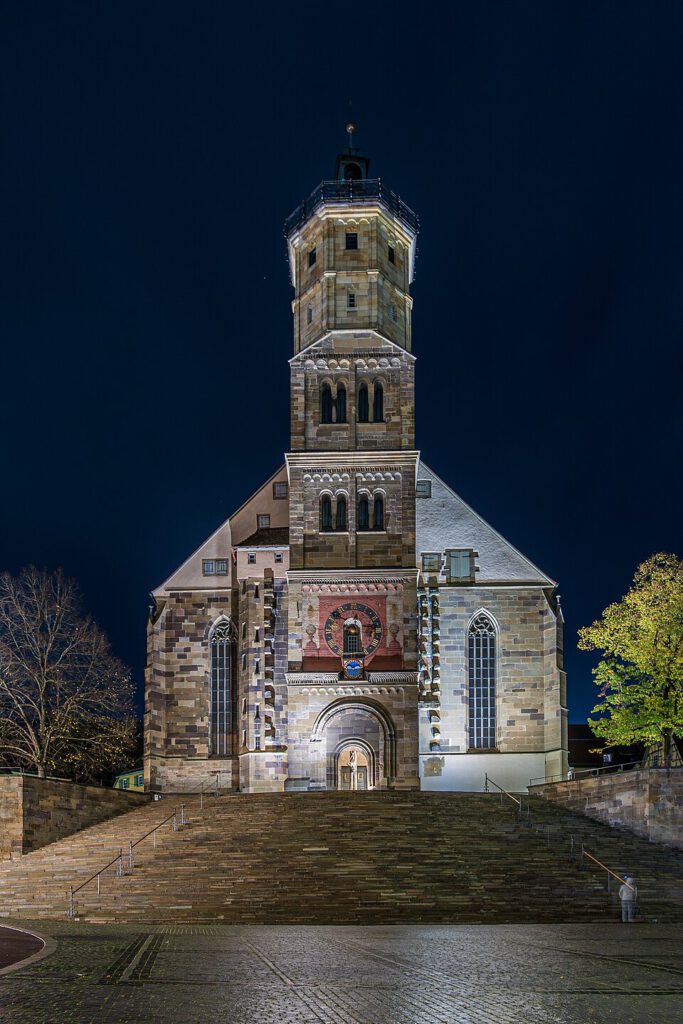 Abendaufnahme der historischen Kirche mit markantem Turm und kunstvollen Fenstern, umgeben von beleuchteten Bäumen und einer steinernen Treppe.