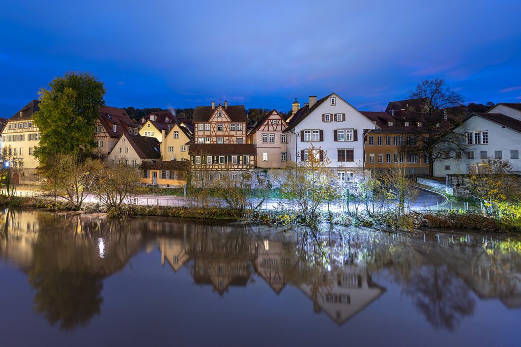 Abendliche Stadtansicht mit historischen Fachwerkhäusern am Ufer eines ruhigen Gewässers, beleuchtet von sanftem Licht unter einem blauen Himmel.