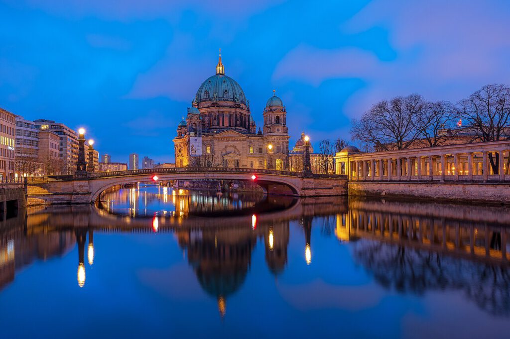 Berliner Dom bei Dämmerung, reflektiert im Wasser des Spreeufers, mit der Oberbaumbrücke im Vordergrund und städtischer Architektur im Hintergrund.