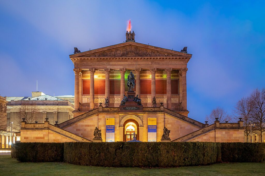 Altes Museum in Berlin bei Nacht, beleuchtet mit bunten Lichtern. Die beeindruckende Fassade zeigt klassische Säulen und eine Statue auf dem Dach. Im Vordergrund sind die Treppen und kunstvoll geschnittene Hecken zu sehen.