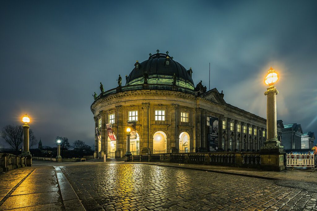 Historisches Gebäude des Bode-Museums in Berlin bei Nacht, beleuchtet von Straßenlaternen, mit reflektierenden Pflastersteinen im Vordergrund.