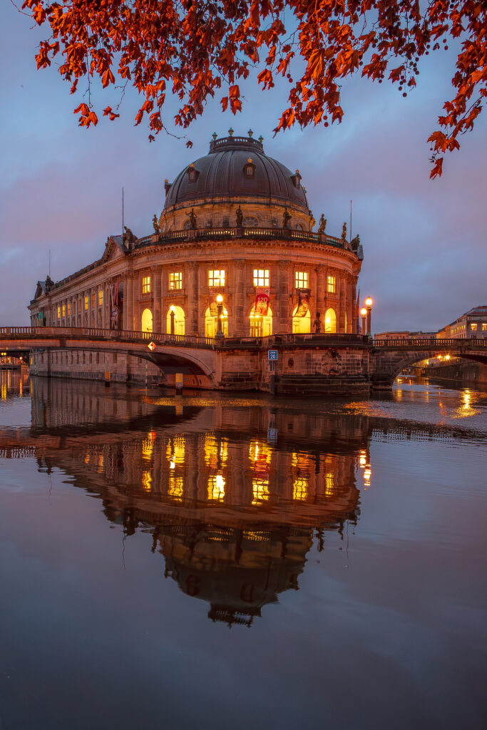 Blick auf das Bode-Museum in Berlin bei Dämmerung, reflektiert im Wasser, umgeben von herbstlichen Blättern. Die warmen Lichter des Museums strahlen eine einladende Atmosphäre aus.