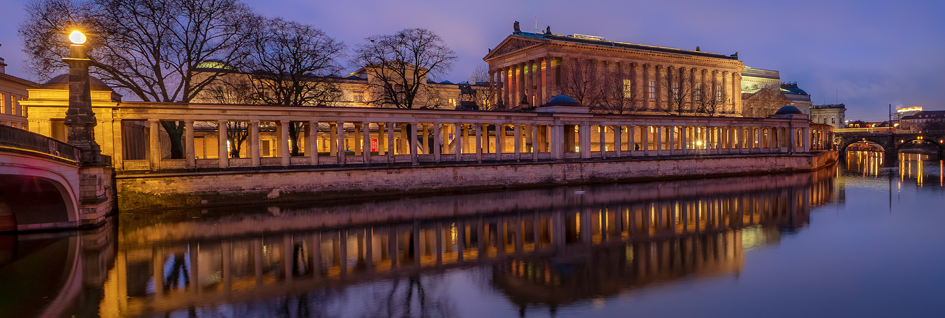 Abendliche Ansicht des Pergamonmuseums in Berlin, reflektiert im Wasser, umgeben von Bäumen und beleuchtet von Straßenlaternen.