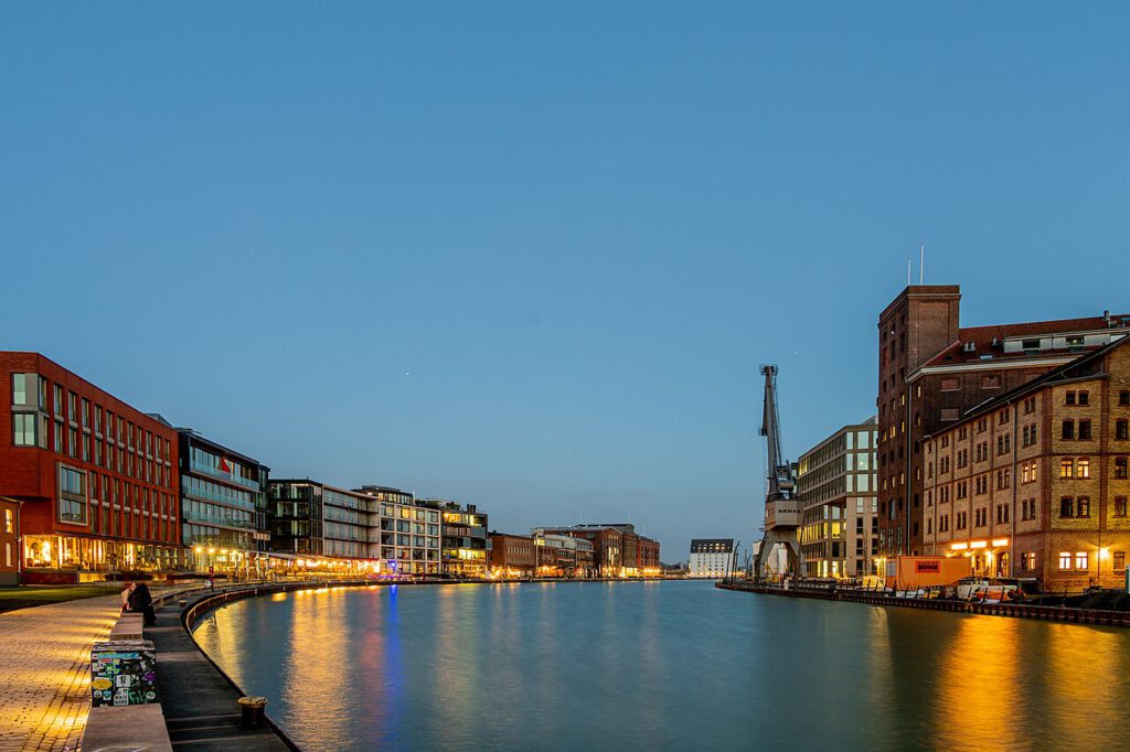 Blick auf den Hafen mit modernen Gebäuden und historischen Lagerhäusern bei Dämmerung, reflektierende Lichter im Wasser.