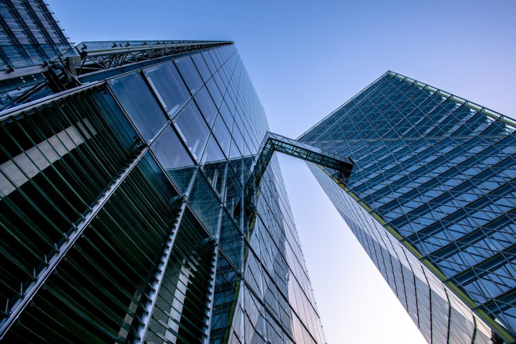 Moderne Architektur von zwei gläsernen Wolkenkratzern, die sich in den Himmel erstrecken und durch eine transparente Brücke verbunden sind. Die Aufnahme zeigt die beeindruckenden Strukturen aus einem tiefen Blickwinkel unter klarem Himmel.