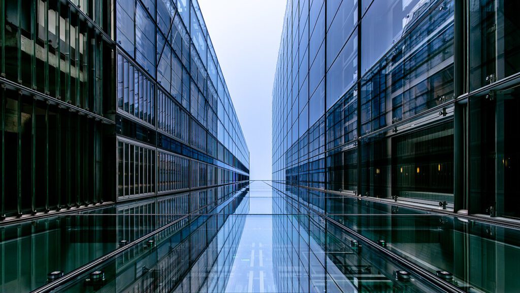 Moderne Architektur mit gläsernen Fassaden, die einen Blick auf den schmalen Durchgang zwischen zwei Gebäuden bietet. Reflexionen auf dem Glas und der klare Himmel schaffen eine beeindruckende Perspektive.