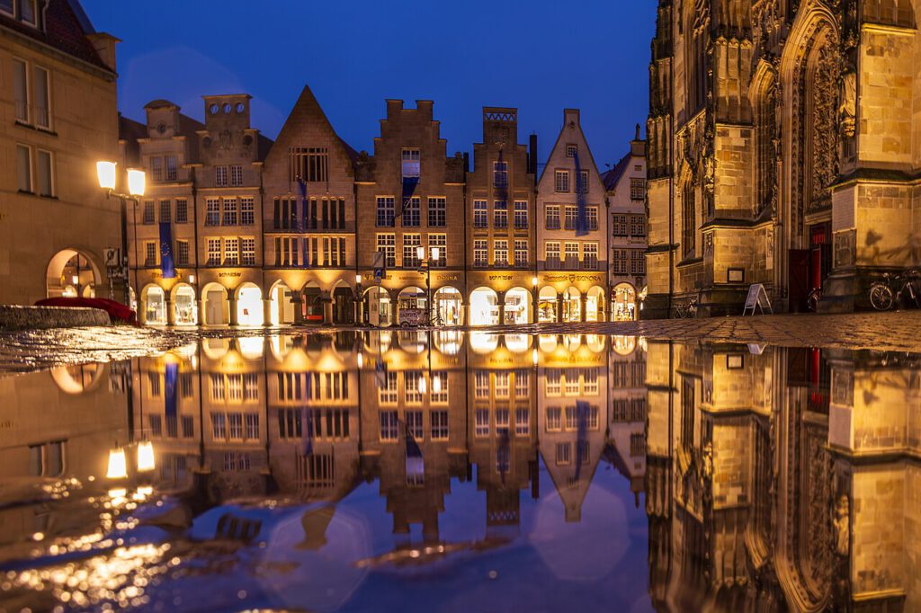 Nachtaufnahme des historischen Marktplatzes in Münster mit beleuchteten Giebelhäusern und reflektierenden Pfützen auf dem Boden.