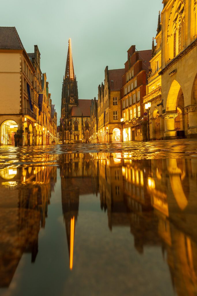 Blick auf die historische Altstadt mit dem St.-Lamberti-Kirchturm, reflektiert im Wasser. Die beleuchteten Gebäude und die stimmungsvolle Atmosphäre schaffen ein malerisches Stadtbild in Münster bei Dämmerung.