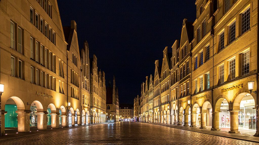 Nachtaufnahme der historischen Altstadt von Münster mit beleuchteten Gebäuden und Arkaden, die eine einladende Atmosphäre schaffen. Die gepflasterte Straße ist menschenleer und bietet einen Blick auf die architektonische Schönheit der Stadt.
