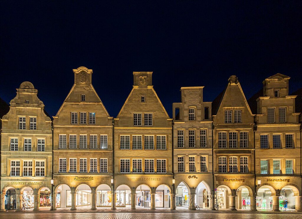 Historische Gebäude am Marktplatz in Münster bei Nacht, mit erleuchteten Schaufenstern und typischer norddeutscher Architektur.