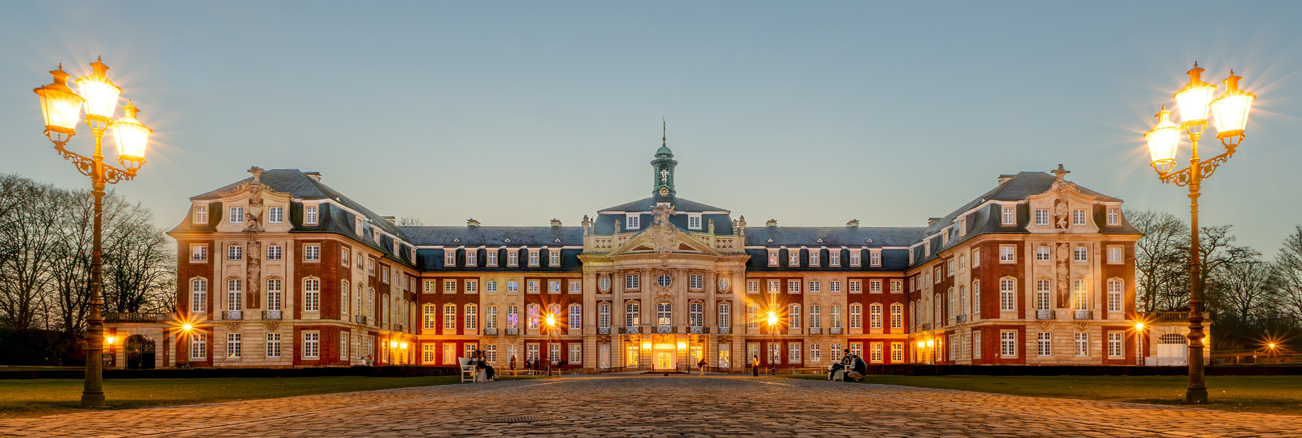 Historisches Schloss mit prächtiger Fassade und beleuchteten Laternen im Vordergrund, umgeben von einem gepflegten Garten bei Dämmerung.