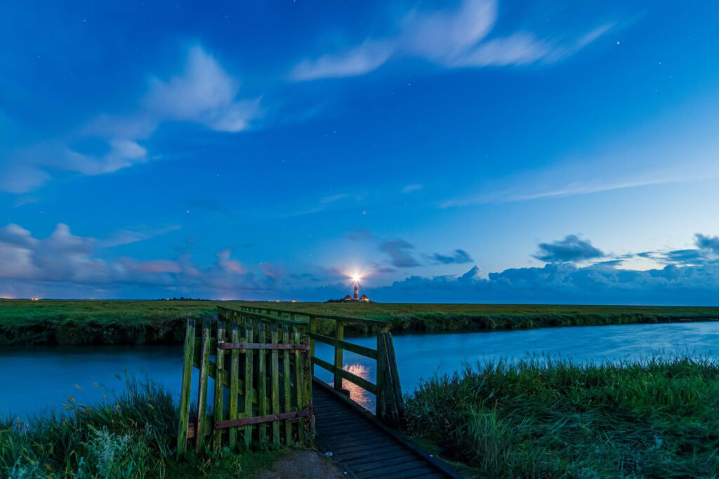 Landschaft bei Nacht mit einem Holzsteg, der zu einem Leuchtturm führt. Der Himmel ist klar und zeigt eine Vielzahl von Wolken und Sternen. Die ruhige Wasseroberfläche reflektiert das Licht des Leuchtturms. Ideal für Natur- und Landschaftsfotografie.