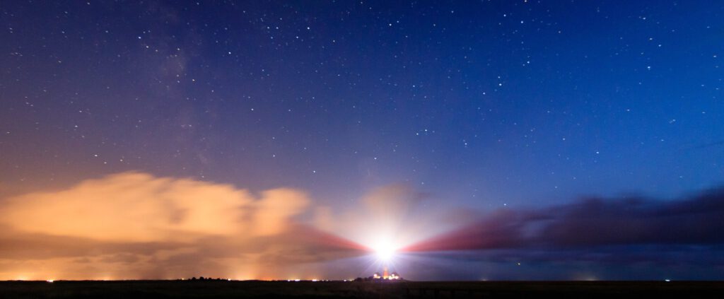 Leuchtturm im nächtlichen Himmel mit Sternen und farbigen Wolken, die von der Lichtquelle des Leuchtturms beleuchtet werden.