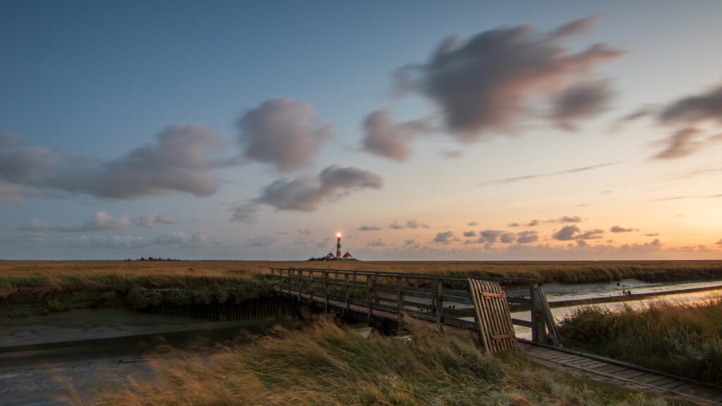 Blick auf einen Leuchtturm im Nationalpark Wattenmeer bei Sonnenuntergang, umgeben von weiten Graslandschaften und einem Holzsteg, der zum Wasser führt.