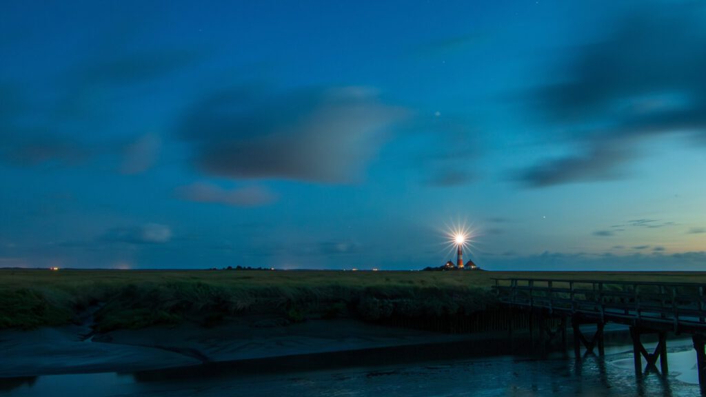 Leuchtturm bei Nacht mit strahlendem Licht, umgeben von grünen Wiesen und einem ruhigen Wasserlauf, unter einem klaren, blauen Himmel.