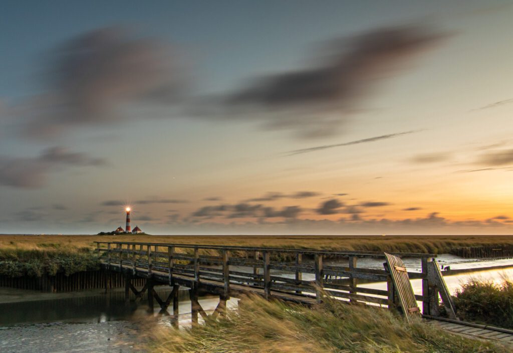 Ein Holzsteg führt zu einem Leuchtturm in der Nordsee, umgeben von grünem Gras und sanften Wellen. Der Sonnenuntergang färbt den Himmel in warmen Tönen und schafft eine ruhige Atmosphäre.