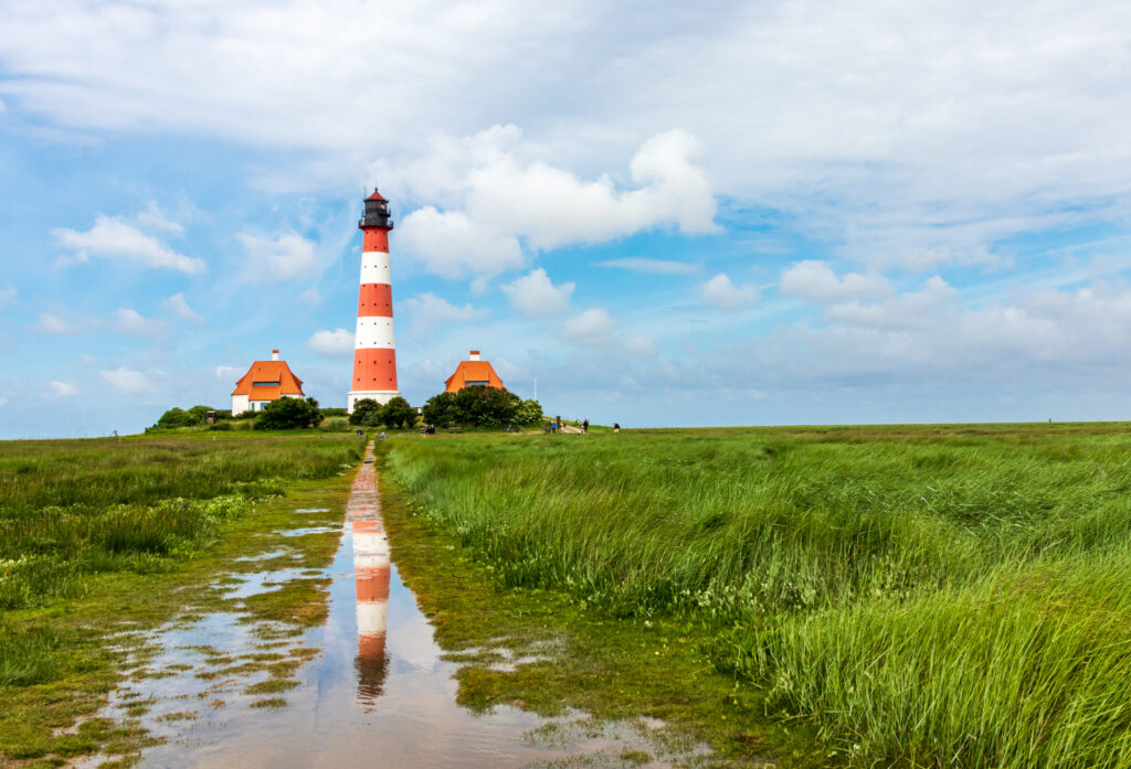 Leuchtturm in St. Peter-Ording mit rot-weißer Streifenmuster, umgeben von grünen Wiesen und einem spiegelnden Wasserweg. Ideal für Fotografien und Erkundungen der Nordsee.