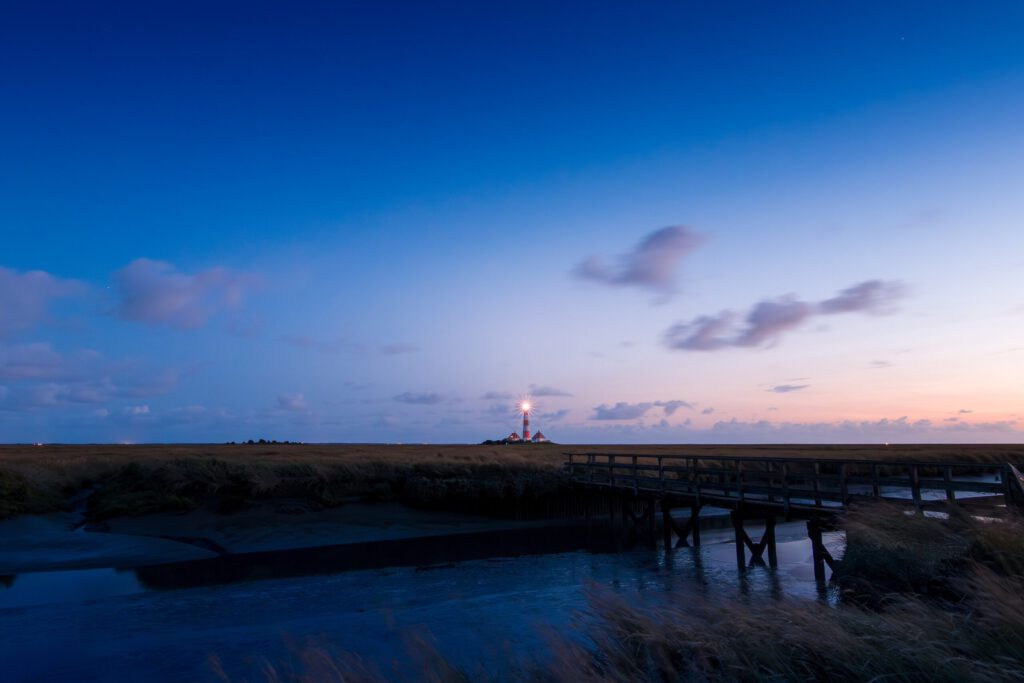 Leuchtturm in der Dämmerung mit Holzsteg und weiten Graslandschaften, die von sanften Wellen umgeben sind.