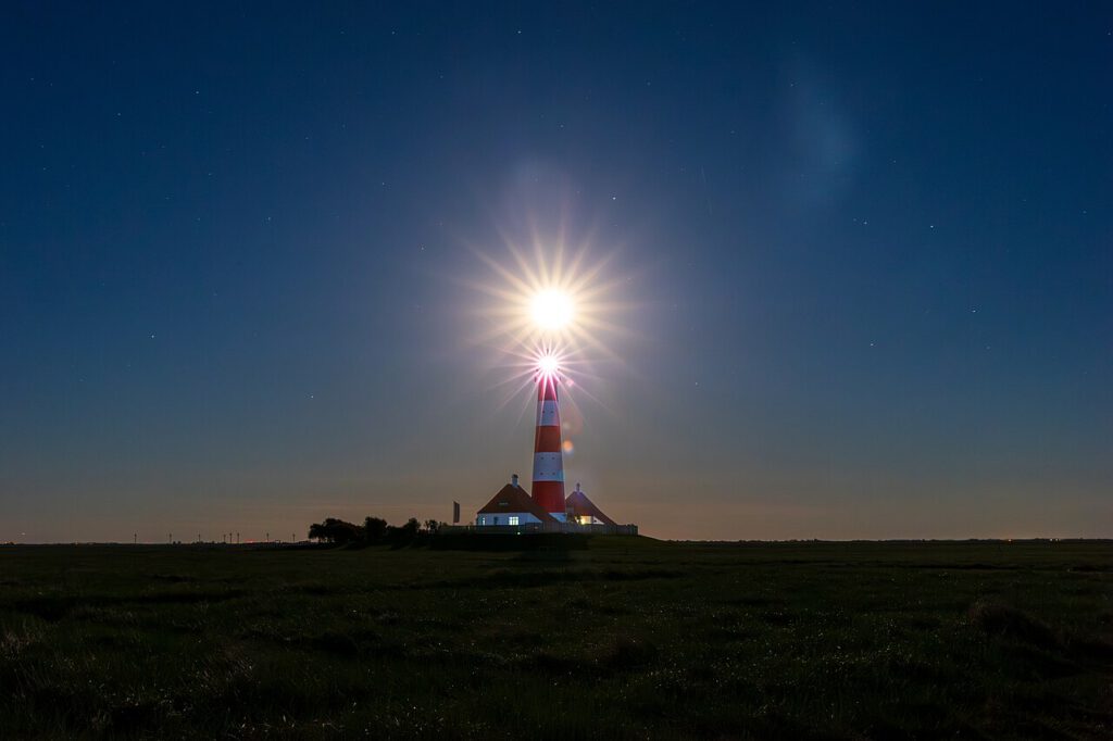 Leuchtturm bei Nacht mit strahlendem Mond und klaren Sternen am Himmel, umgeben von grünem Grasland.