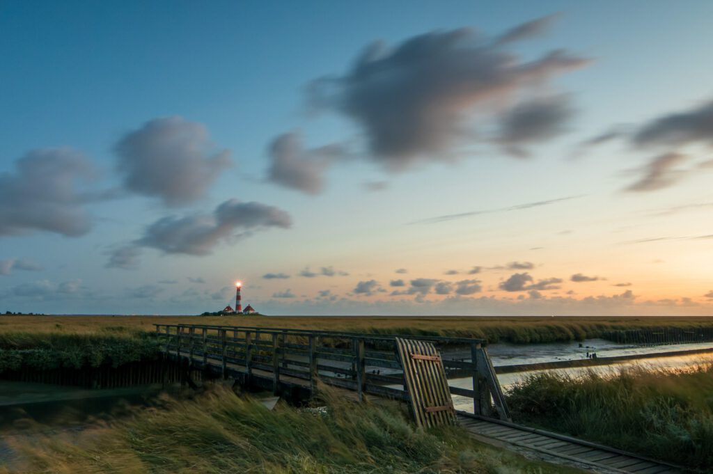 Leuchtturm bei Sonnenuntergang in Norddeutschland, umgeben von Wiesen und Wolken, mit einem Holzsteg im Vordergrund. Ideal für Natur- und Landschaftsfotografie.