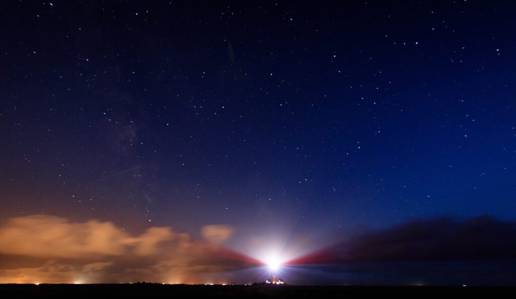 Leuchtturm im nächtlichen Himmel mit Sternen und Wolken, beleuchtet von einem starken Lichtstrahl.