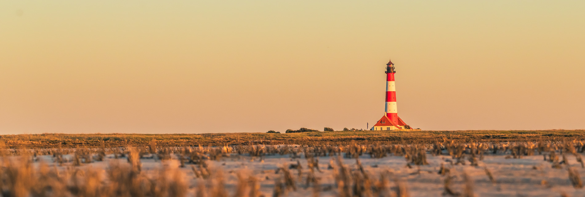 Leuchtturm auf der Nordseeinsel Amrum bei Sonnenuntergang, umgeben von weiten Wattlandschaften und sanften Farben des Himmels.
