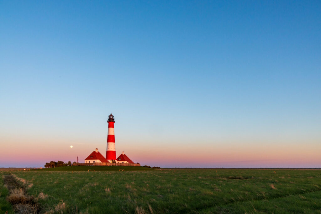 Leuchtturm von Westerhever bei Sonnenuntergang, umgeben von grünen Wiesen und klarem Himmel, mit Vollmond im Hintergrund.
