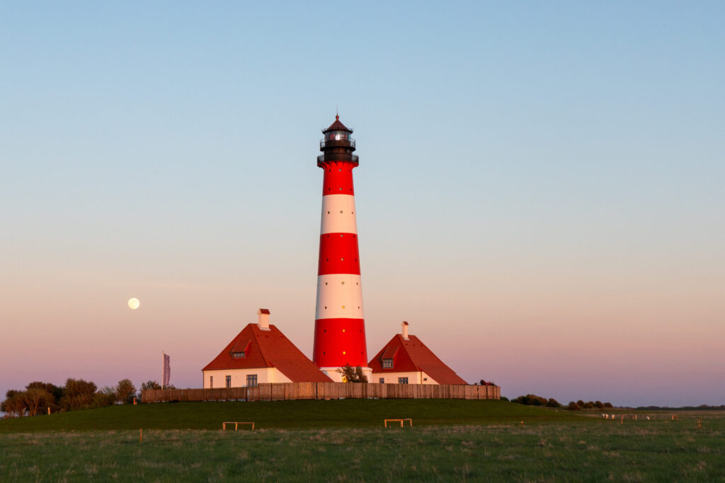 Leuchtturm mit roter und weißer Streifenmuster vor einem sanften Abendhimmel, umgeben von Wiesen und Bäumen, mit dem Mond im Hintergrund.