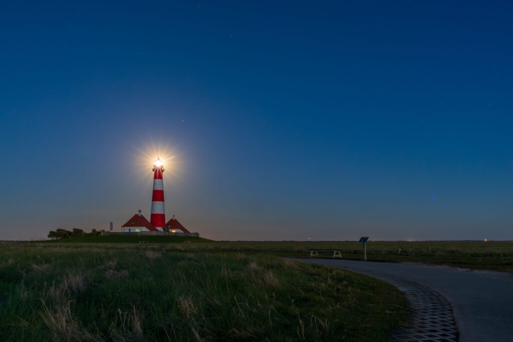 Leuchtturm in der Dämmerung mit strahlendem Licht, umgeben von grünen Wiesen und einem klaren, blauen Himmel. Ideal für maritime Fotografie und Landschaftsaufnahmen.