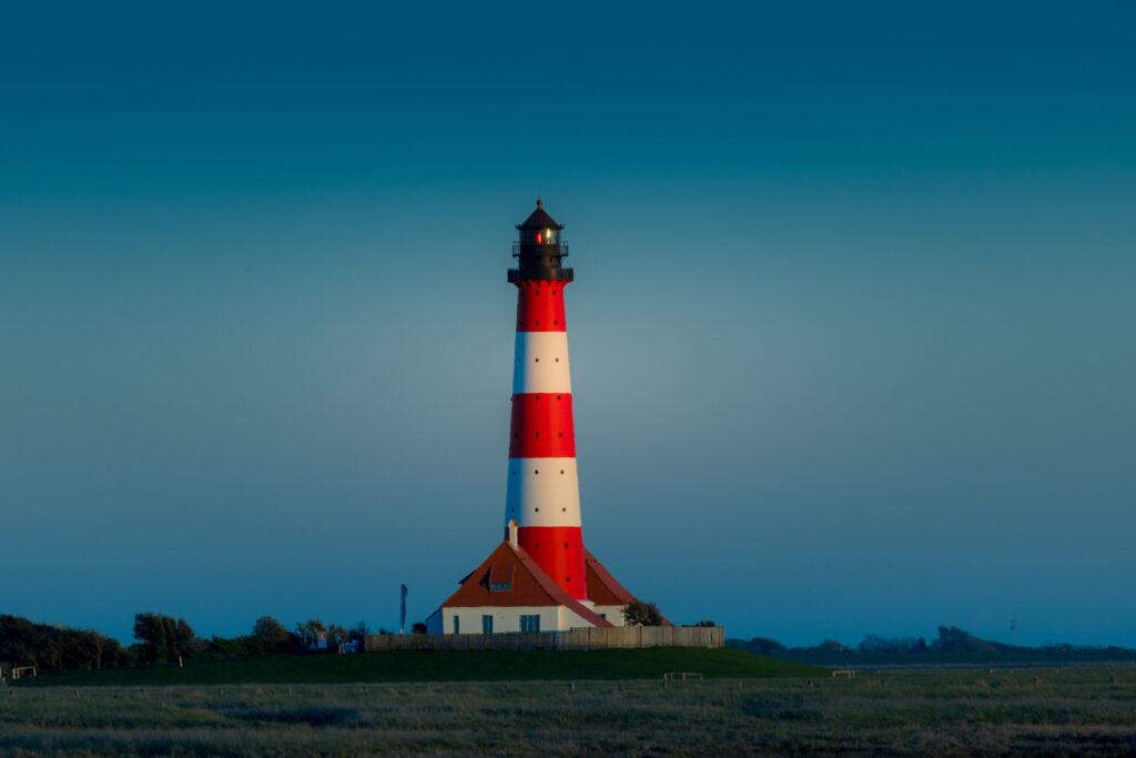 Leuchtturm mit rot-weißen Streifen auf einer grünen Wiese, umgeben von einem klaren blauen Himmel.